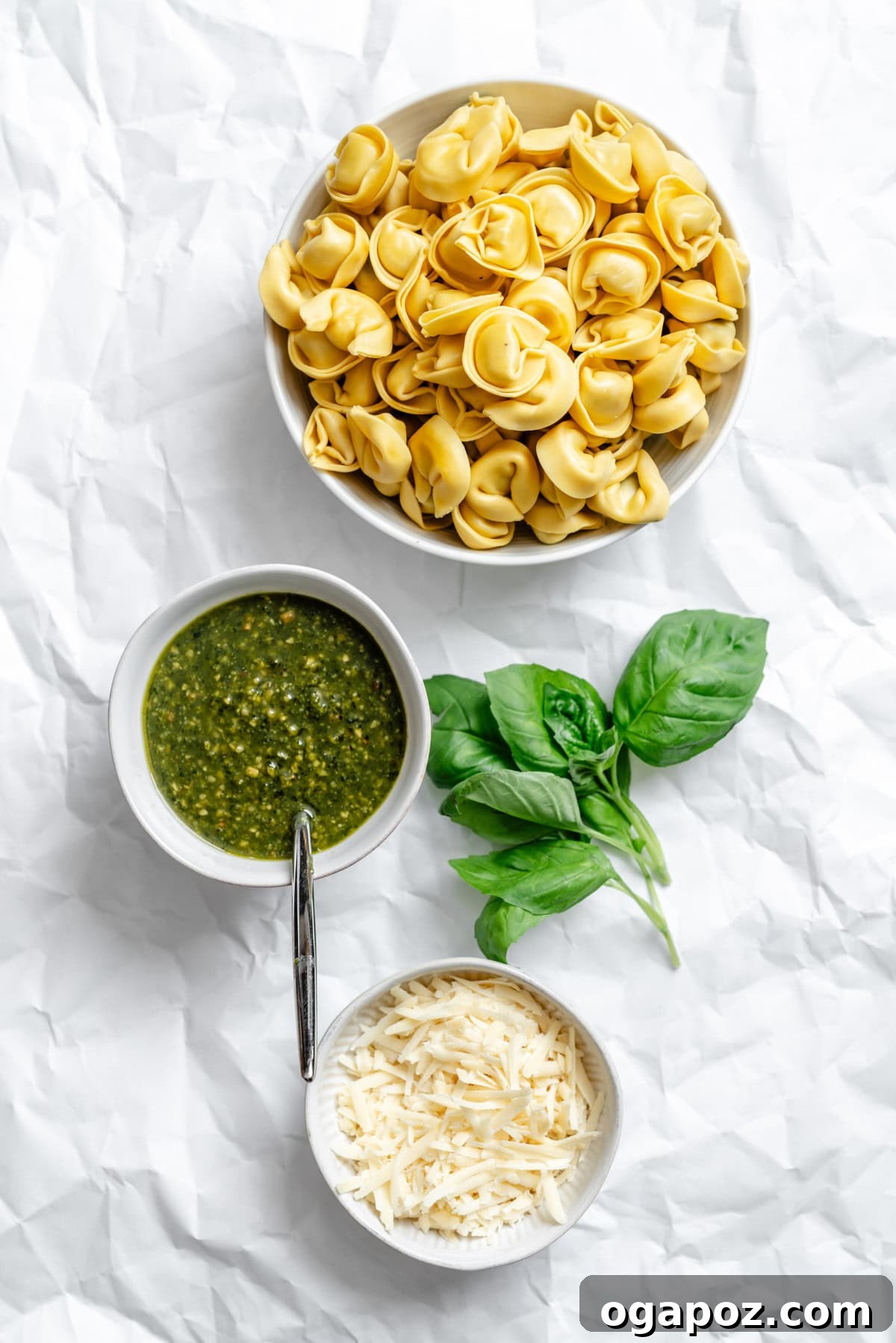 Small bowls containing the main ingredients for pesto tortellini: fresh tortellini, grated Parmesan cheese, and a vibrant green pesto sauce.