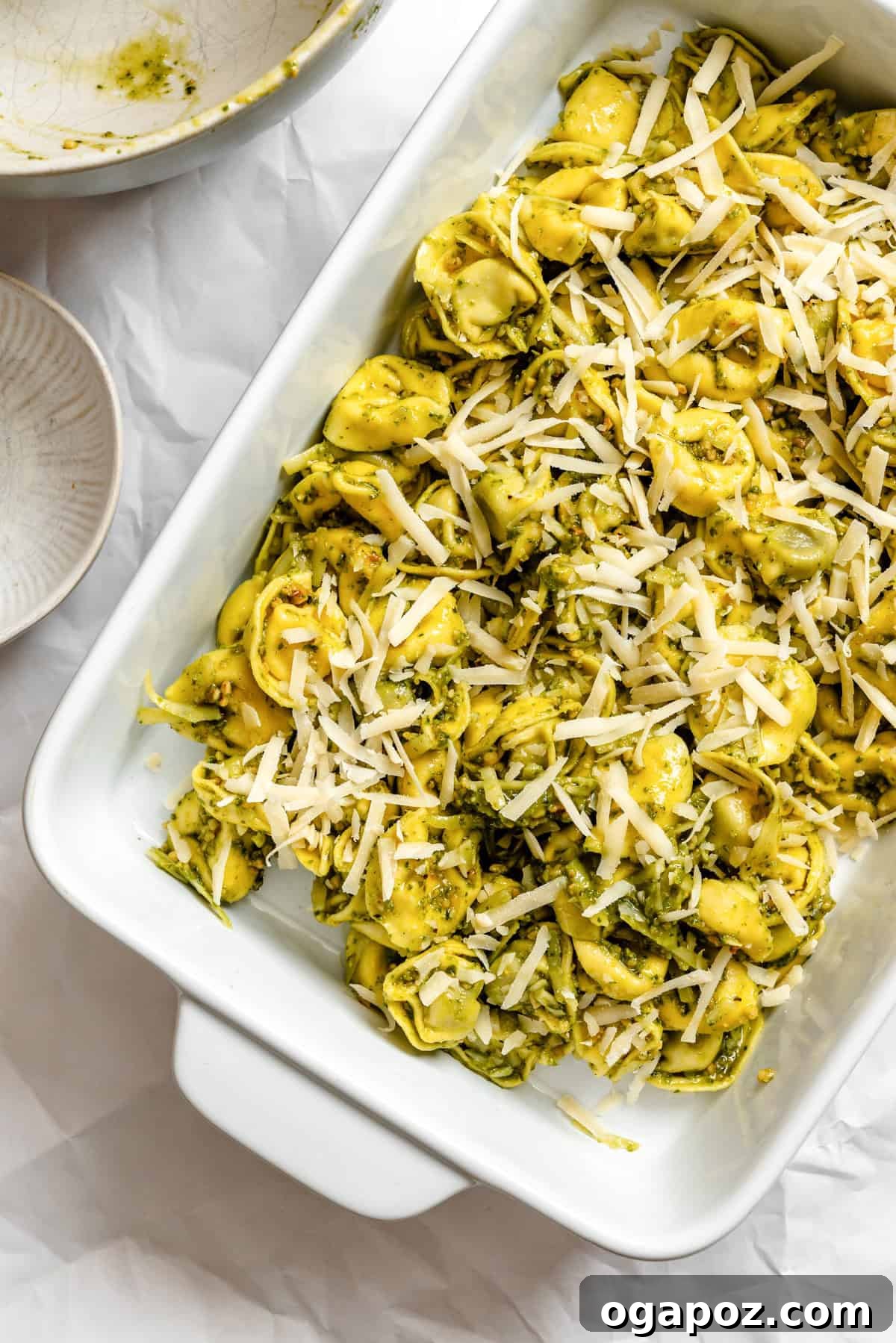 Pesto tortellini spread evenly in a glass baking dish, generously topped with an extra layer of Parmesan cheese before baking.