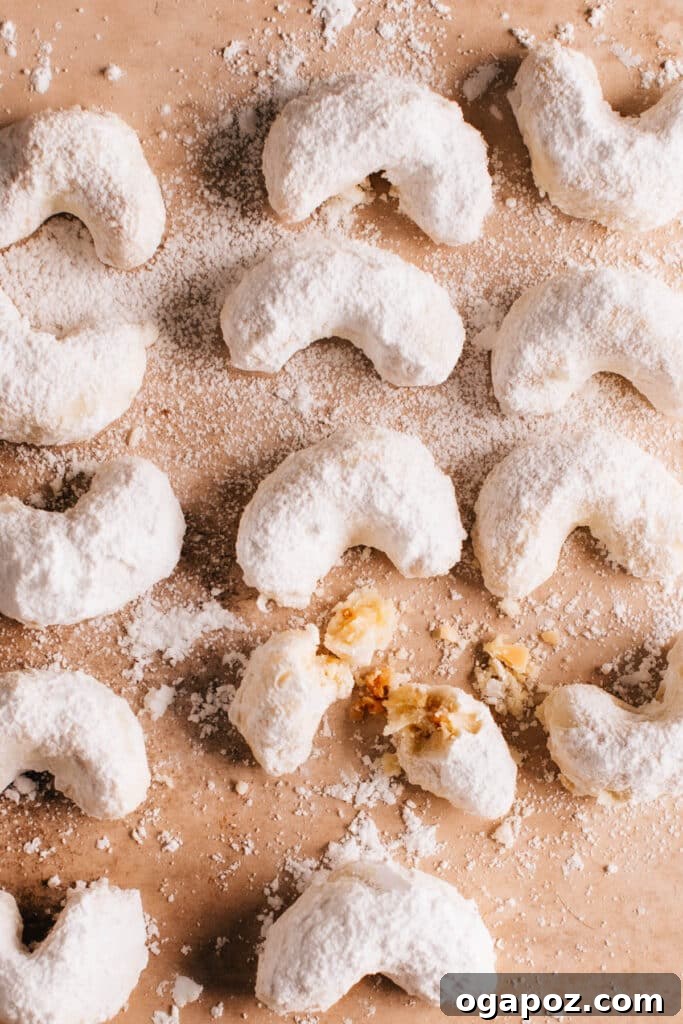 Snow Dusted Kourabiedes A Greek Christmas Classic 2 A beautifully arranged batch of freshly baked Kourabiedes, traditional Greek Christmas cookies, generously dusted with powdered sugar. One cookie is broken in half, revealing its tender, crumbly interior filled with toasted almonds, symbolizing the perfect melt-in-your-mouth texture.