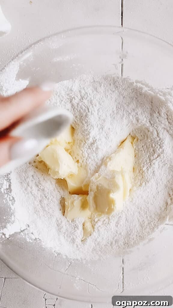 Snow Dusted Kourabiedes A Greek Christmas Classic 4 A hand adding softened butter pieces to a bowl of flour mixture for Kourabiedes dough, showcasing the essential first step of the reverse creaming method for maximum tenderness.