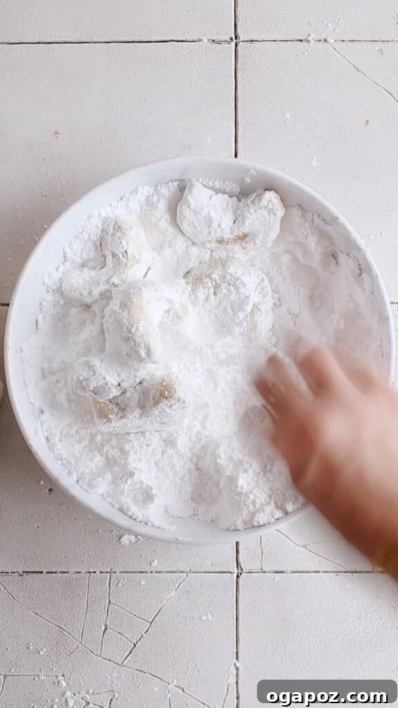 Snow Dusted Kourabiedes A Greek Christmas Classic 10 Close-up photograph of Kourabiedes being fully and generously coated in a thick, snowy layer of powdered sugar within a shallow bowl, clearly demonstrating the thorough and traditional dusting process that gives them their iconic festive appearance.