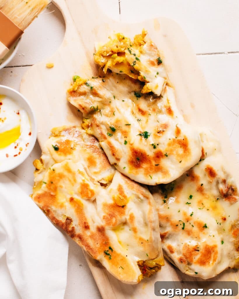 Stuffed Potato Naan 2 Aloo naan served with dip, one naan torn to reveal filling of potato masala, on a wooden chopping board.