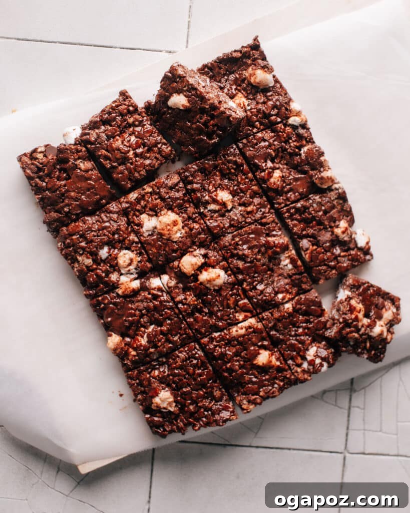 Chocolate rice krispie treats are cut into even squares on a white parchment paper, ready to be served. The treats have a gooey texture from the melted marshmallows and chocolate chips.