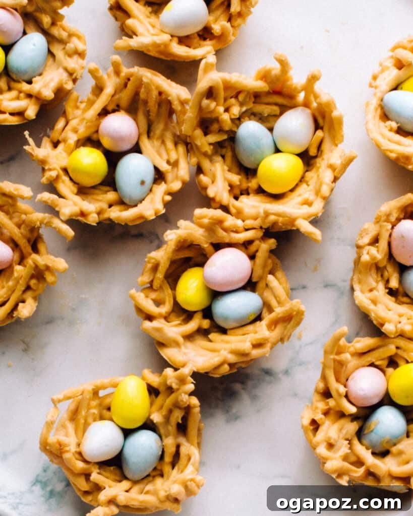 Bird Nest Treats 2 Close-up of three bird nest cookies, each filled with three pastel chocolate Easter eggs, resting in a muffin tin liner. The cookies have a textured, twig-like appearance from the chow mein noodles and a creamy, white chocolate coating.