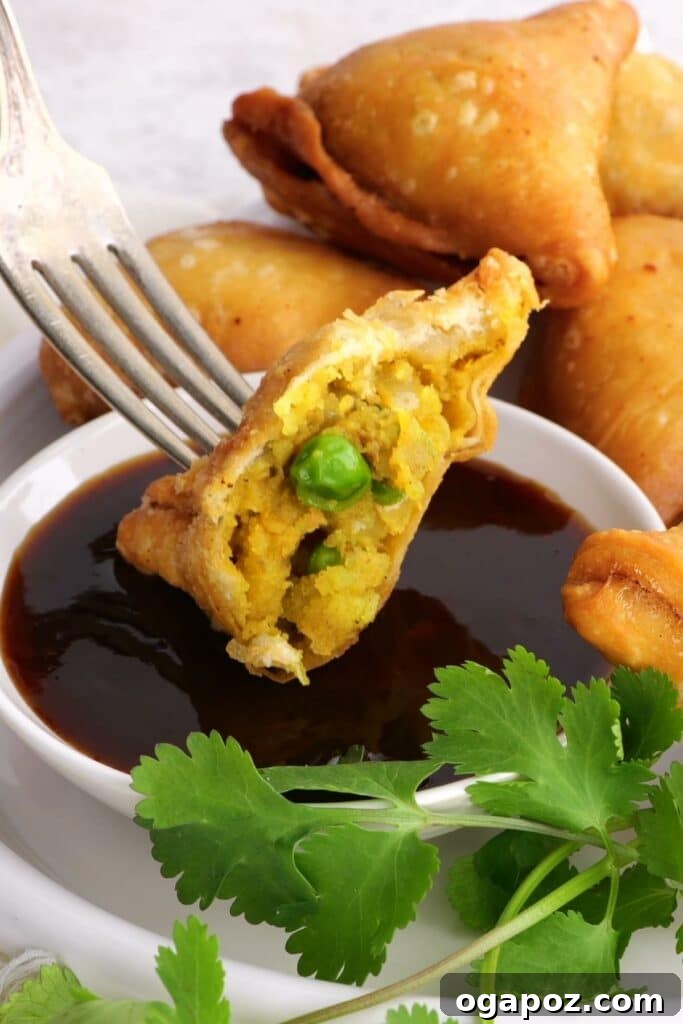 Flaky Spiced Potato Pockets 7 A crisp potato samosa, freshly fried and golden, is shown with a fork taking a bite, dipped in a side of green chutney, highlighting its delicious texture.