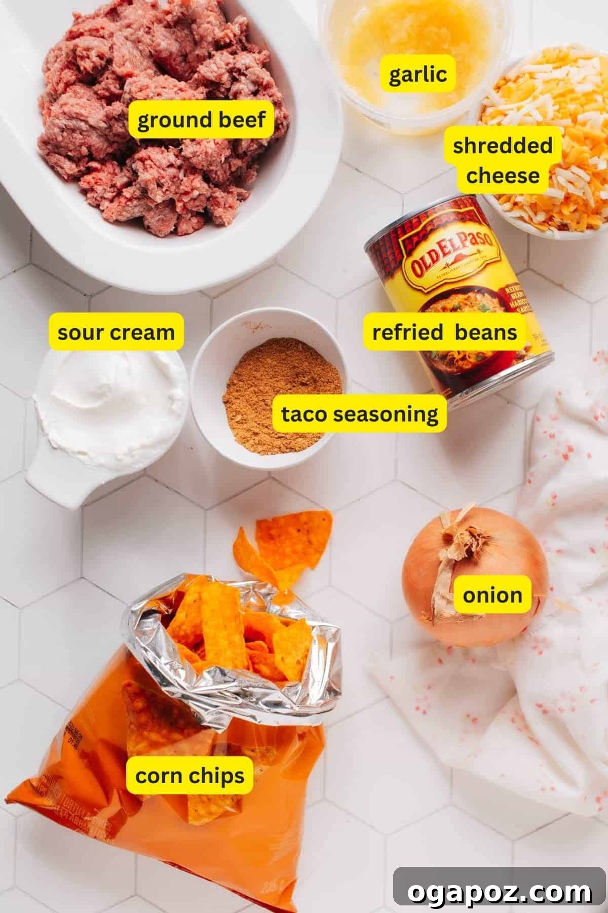 Portable Fiesta Bake 3 A carefully arranged display of ingredients for Walking Taco Casserole, including lean ground beef, refried beans, taco seasoning, sour cream, shredded cheese, a yellow onion, garlic cloves, and a bag of corn chips.