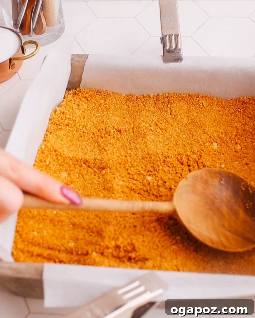 Graham cracker crumbs mixed with melted butter being pressed firmly into the bottom of a square baking pan lined with parchment paper, forming the crust for Pumpkin Cheesecake Bars.