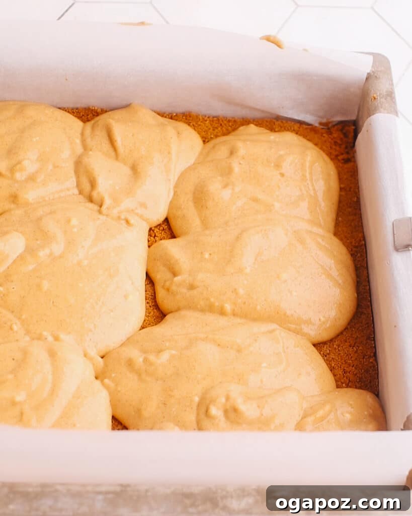 The orange-hued pumpkin cheesecake batter being spread evenly over the pre-baked graham cracker crust in a square baking pan.