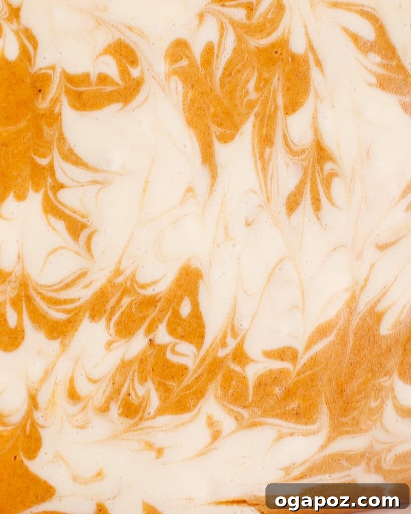 An up-close, textured shot of the beautiful orange and cream marbled swirl of pumpkin and vanilla cheesecake batter before baking, showcasing the intricate pattern.