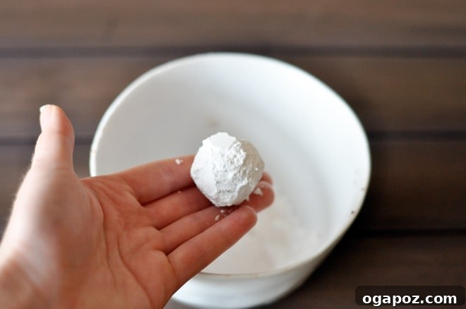 Decadent Chocolate Snowball Cookies 4 A hand rolling chocolate snowball cookie dough in a bowl of powdered sugar, ensuring a thick coating.