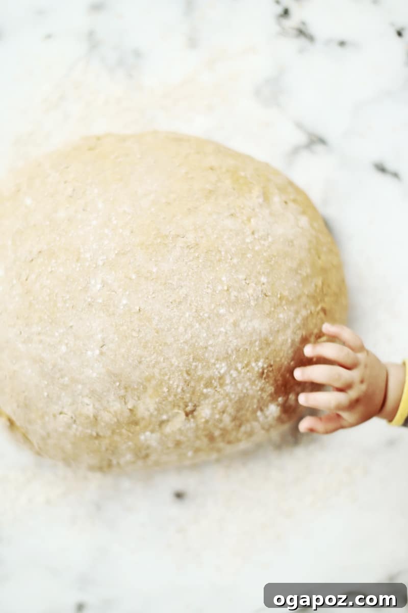 A close-up shot of a small child's chubby fingers gently touching or pulling at the surface of the large, risen bread dough.