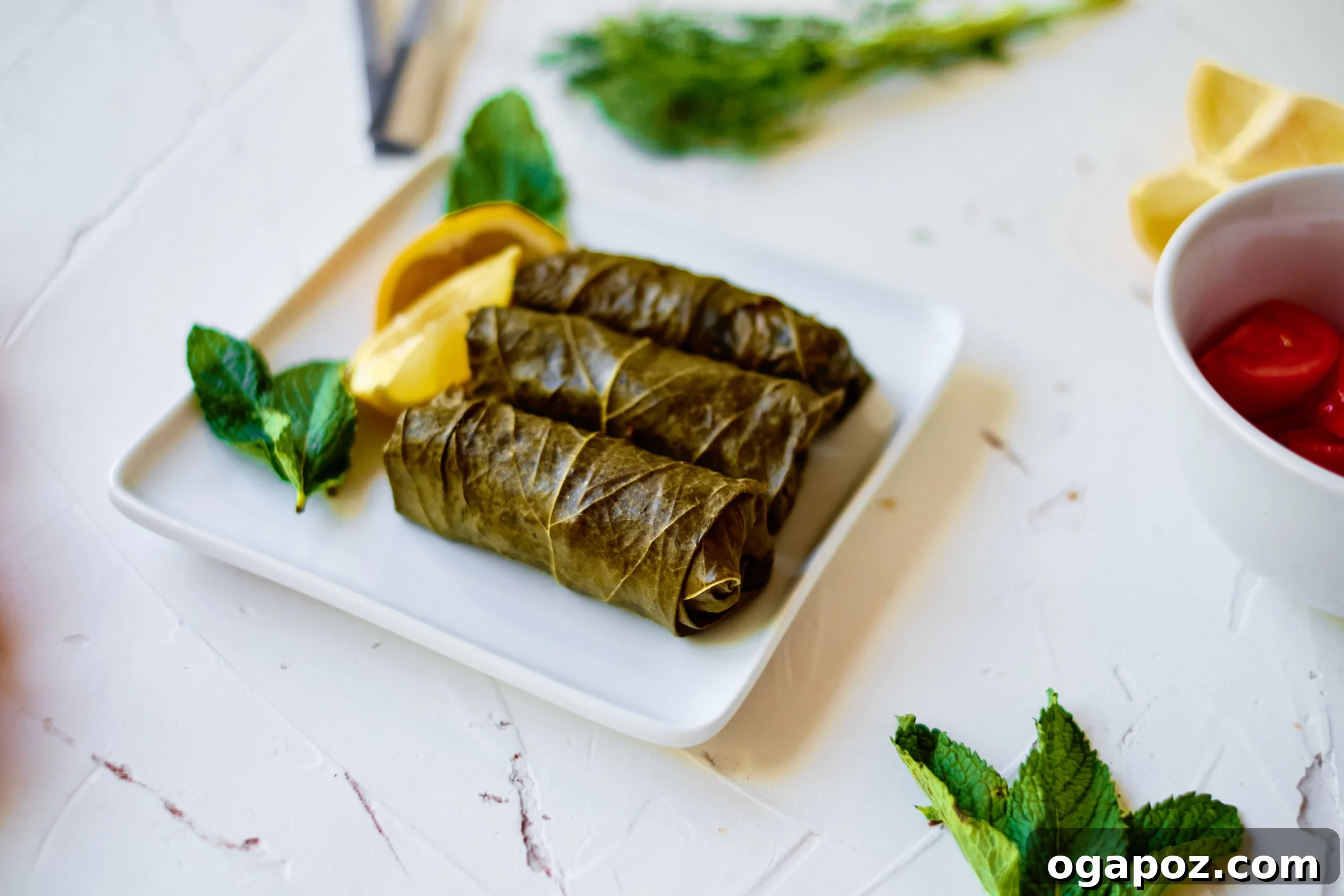 Effortless Dolmades 3 A selection of fresh ingredients for stuffed grape leaves, including uncooked rice, toasted pine nuts, fresh mint, dill, and a jar of grape leaves, arranged on a white cutting board.
