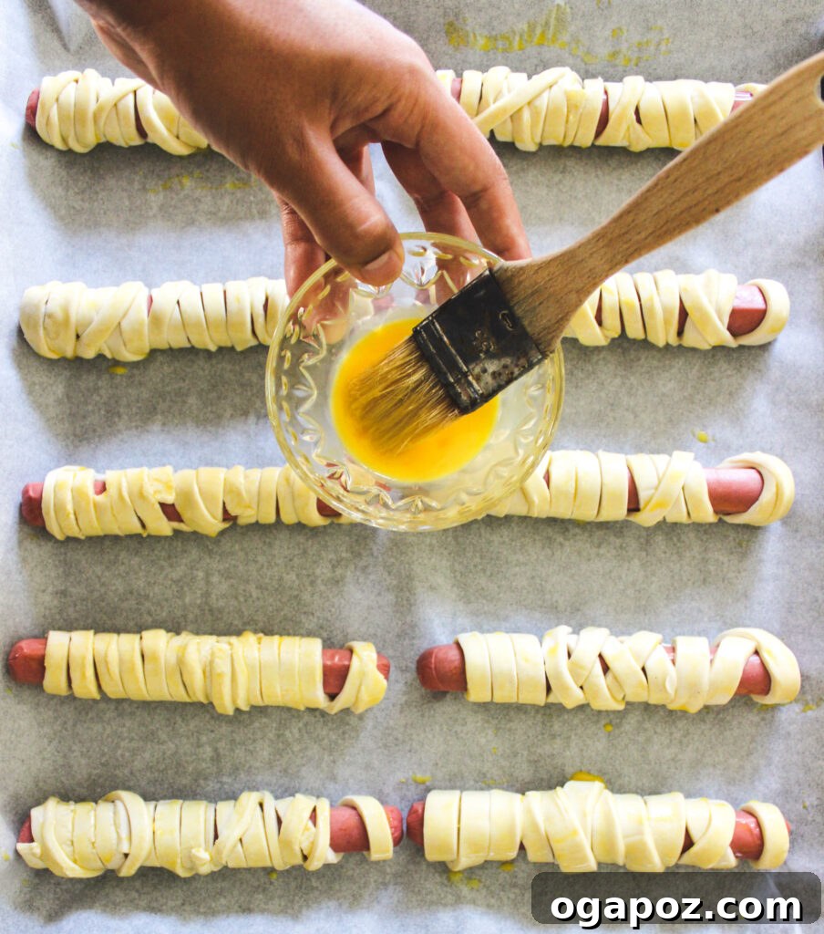 Spooky Sausage Wraps 6 Mummy Hot Dogs on a baking sheet, brushed with egg wash and ready for the oven.