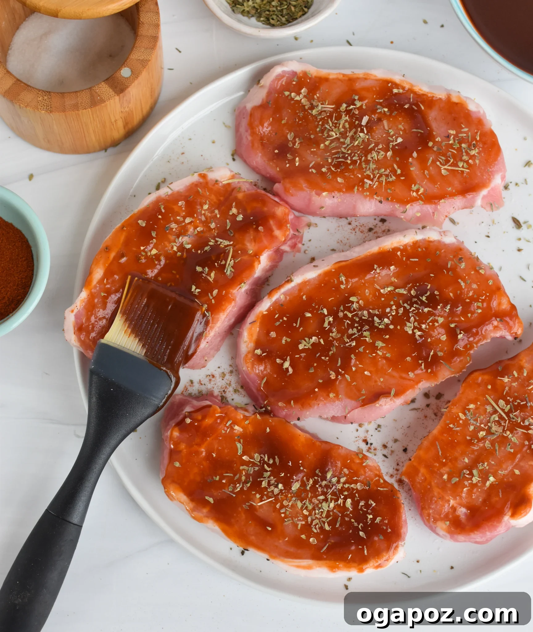 Succulent Oven Baked BBQ Pork Chops 6 A hand brushing BBQ sauce onto a raw pork chop, with a bowl of sauce and a brush nearby.