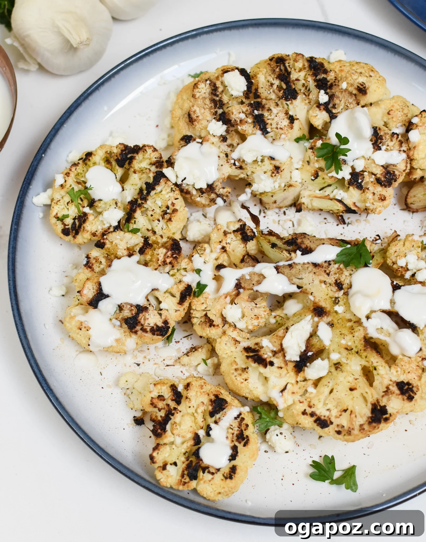 Smoky Grilled Cauliflower Steaks with Creamy Tahini Yogurt 2 Grilled Cauliflower Steaks with Yogurt Tahini Sauce served on a white plate, garnished with fresh herbs and feta cheese.