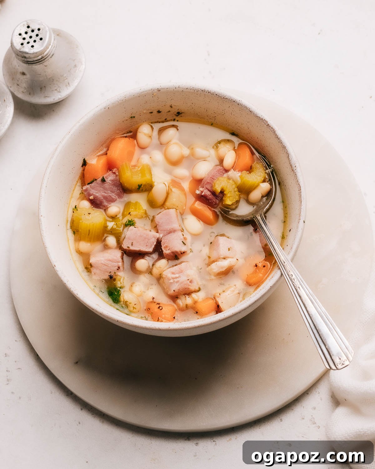 Deliciously Simple Ham Bone Soup Hearty and Flavorful 9 A hearty bowl of Ham Bone Soup with a spoon, alongside salt and pepper shakers on a rustic wooden surface.
