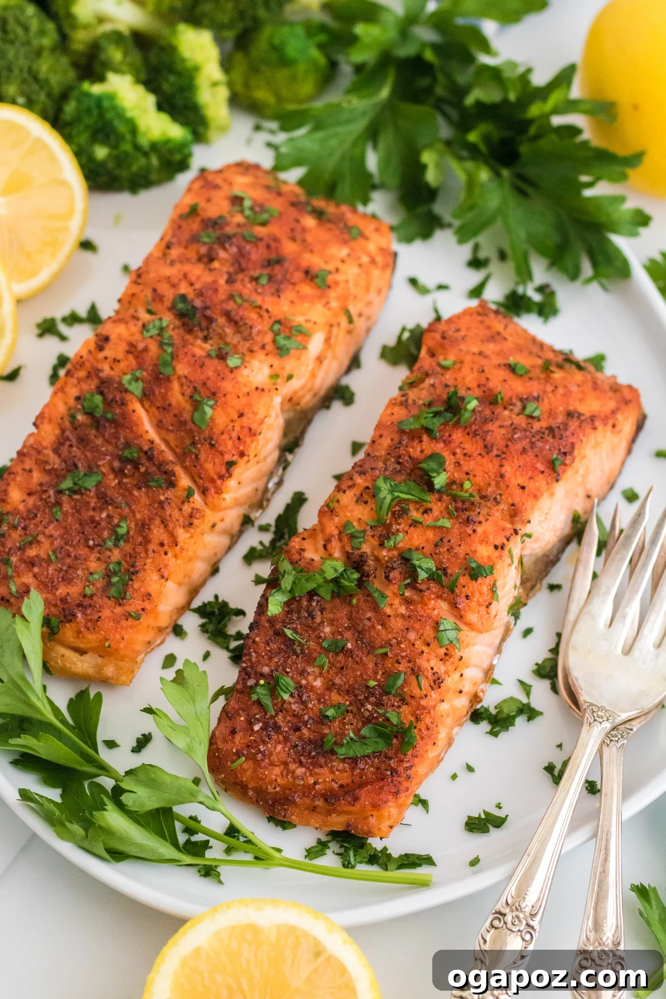 Simple Air Fryer Salmon Perfection 2 Two seasoned and cooked salmon fillets garnished with chopped herbs are served on a white plate. Sliced lemons, parsley leaves, and broccoli florets surround the fillets. Two vintage-style forks are placed beside the salmon.
