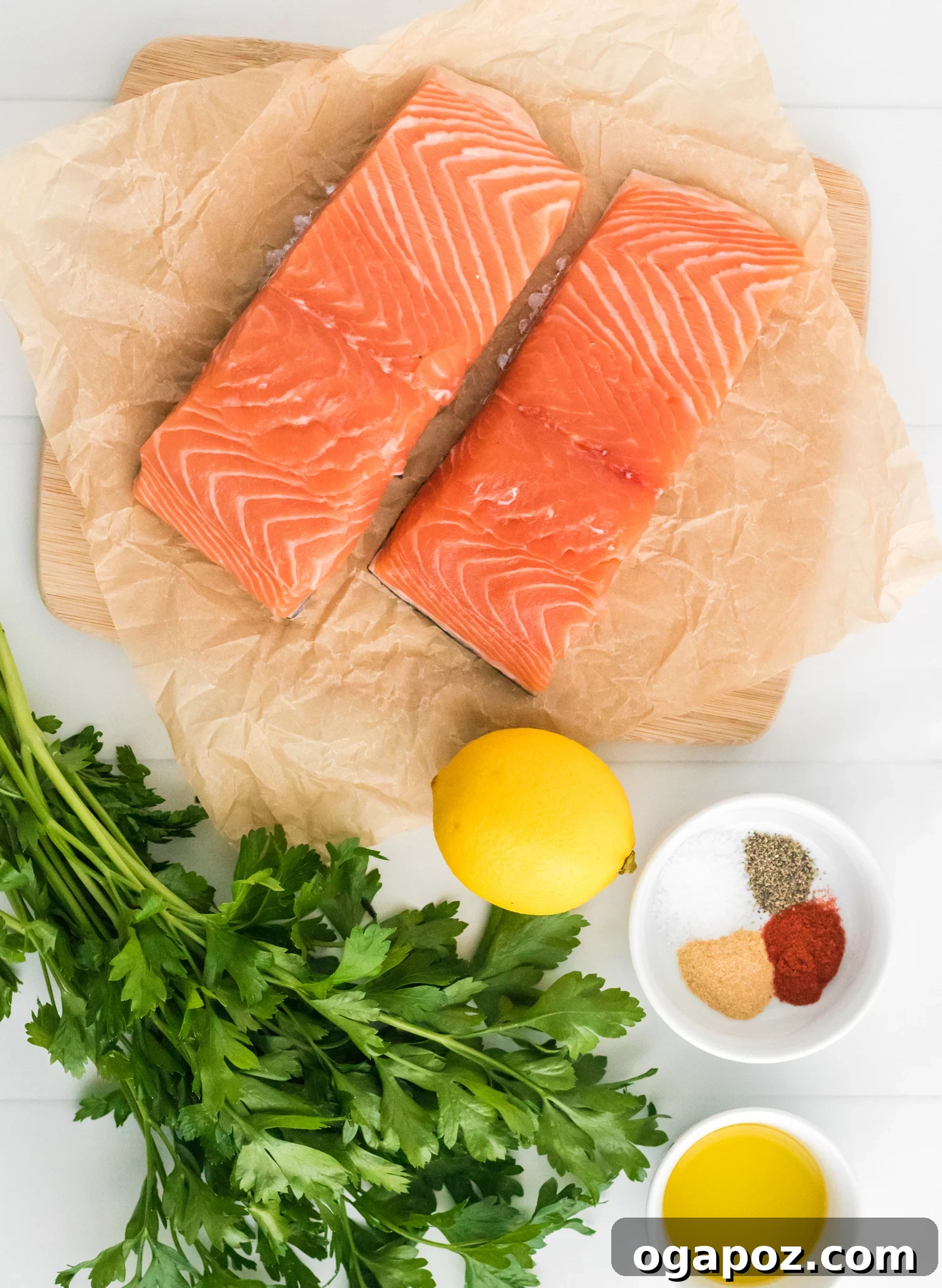 Simple Air Fryer Salmon Perfection 3 Two raw salmon fillets on parchment paper atop a wooden board. A bunch of fresh parsley, a lemon, and a small bowl containing spices, salt, and pepper are beside a bowl of olive oil on a white surface.