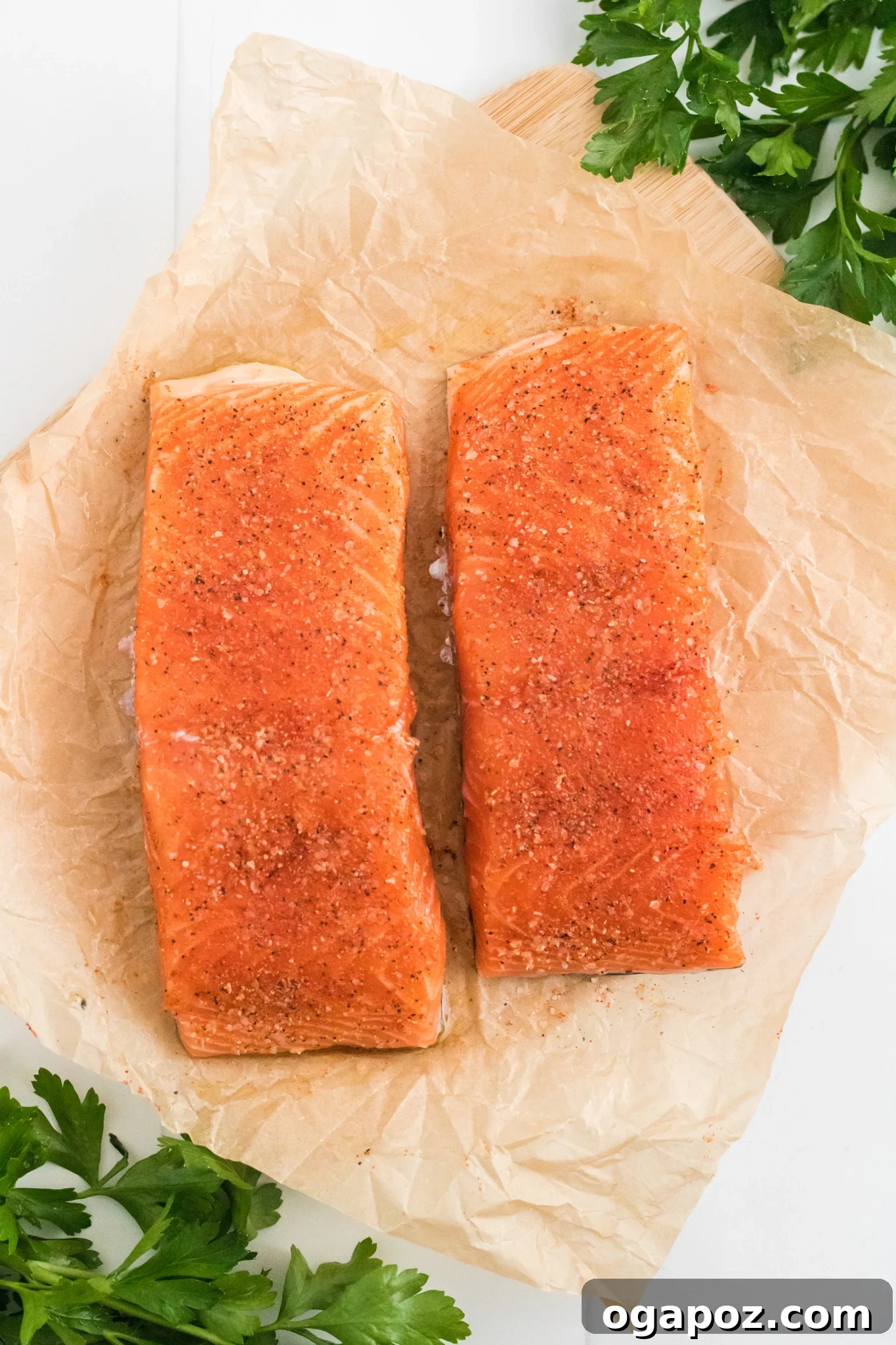 Simple Air Fryer Salmon Perfection 4 Two raw salmon fillets seasoned with spices rest on crinkled parchment paper. Fresh parsley leaves are scattered around, adding a touch of green to the image. The setup is on a light-colored surface.