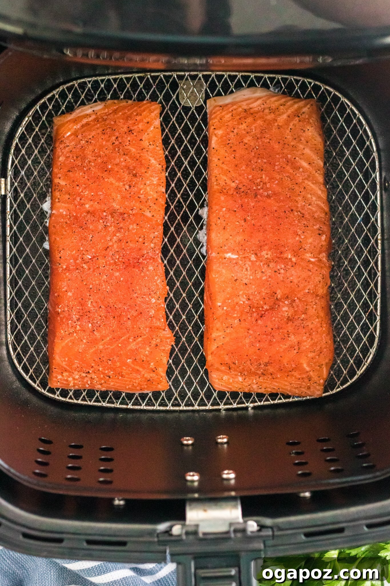Simple Air Fryer Salmon Perfection 5 Two seasoned salmon fillets are placed side by side on the grill rack of an air fryer, ready for cooking. Fresh parsley and a blue-striped cloth can be seen below the open fryer basket.