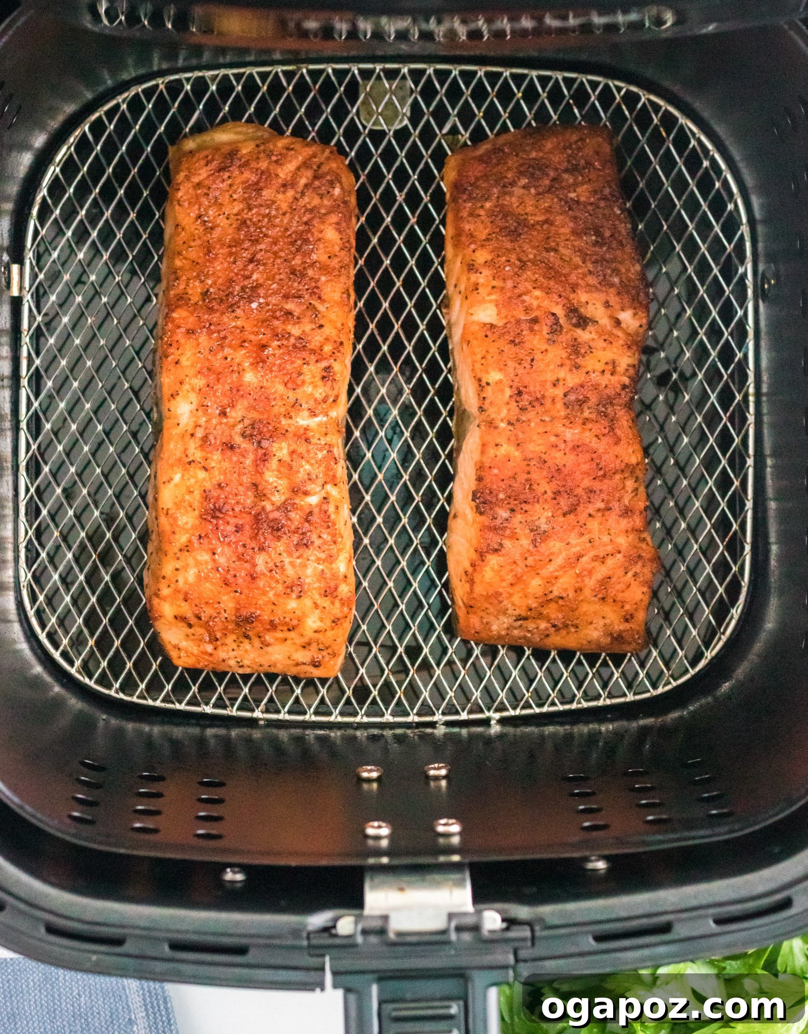 Simple Air Fryer Salmon Perfection 6 Two seasoned salmon fillets are cooking in an air fryer basket. The fillets are golden brown with a crispy texture. Fresh herbs are visible in the bottom right corner.