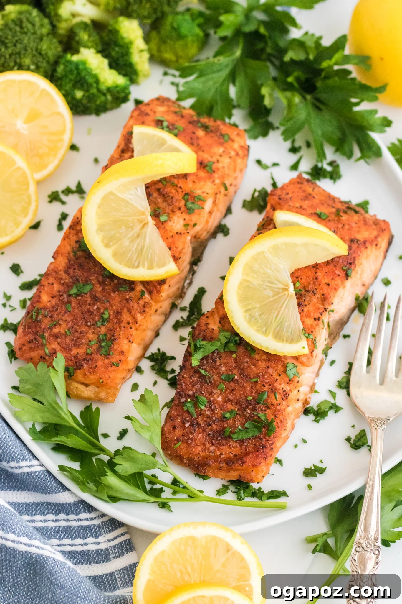 Simple Air Fryer Salmon Perfection 7 Two seasoned salmon fillets on a white plate, each garnished with a lemon slice and chopped herbs. Surrounding the salmon are sprigs of parsley and lemon wedges. A fork and broccoli are visible next to the plate.