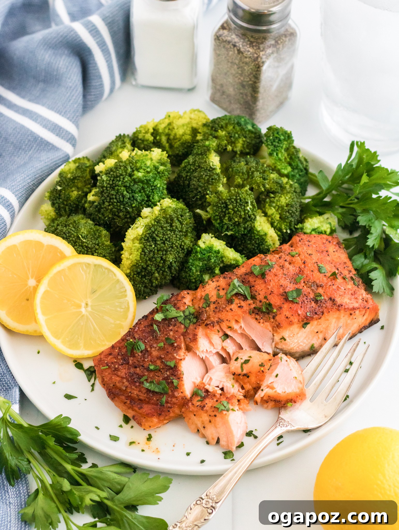 Simple Air Fryer Salmon Perfection 8 A plate with roasted salmon garnished with herbs, steamed broccoli, and lemon slices. A fork flakes the salmon. Salt and pepper shakers, a glass of water, and a striped cloth are in the background. Fresh parsley and a lemon are nearby.