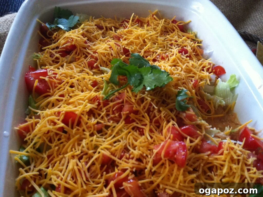 Healthy Taco Dip Close up shot of a vibrant Skinny Seven Layer Dip in a clear dish, showing the distinct layers of beans, cream sauce, salsa, lettuce, tomatoes, cheese, and a sprinkle of cilantro.
