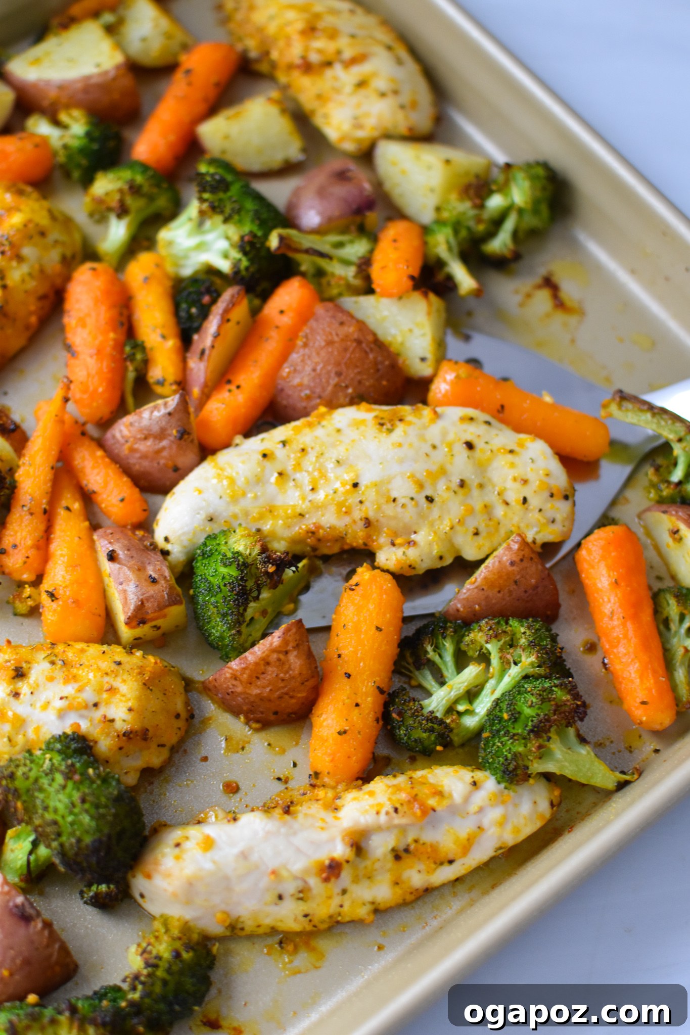 Tek Tepside Limonlu Parmesanlı Tavuk 10 Freshly baked Sheet Pan Lemon Parmesan Chicken Tenders with roasted vegetables on a baking sheet, being scooped with a spatula.