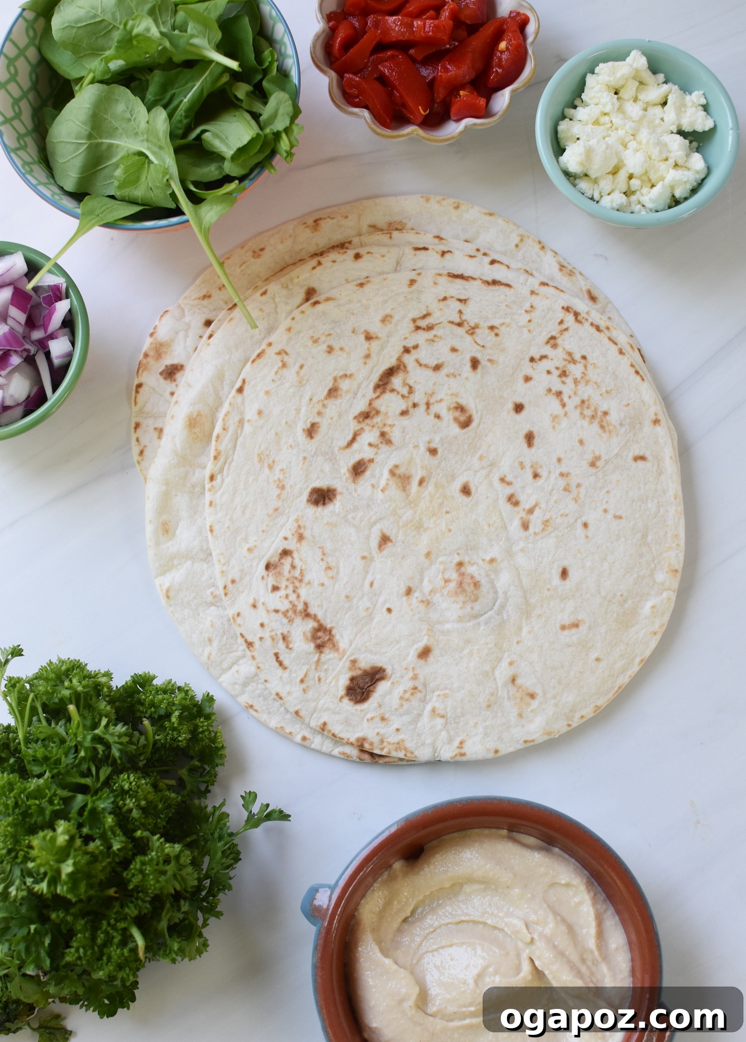 All the fresh ingredients for Mediterranean Pinwheels laid out on a kitchen counter, including tortillas, hummus, feta, roasted red peppers, arugula, red onion, and parsley.