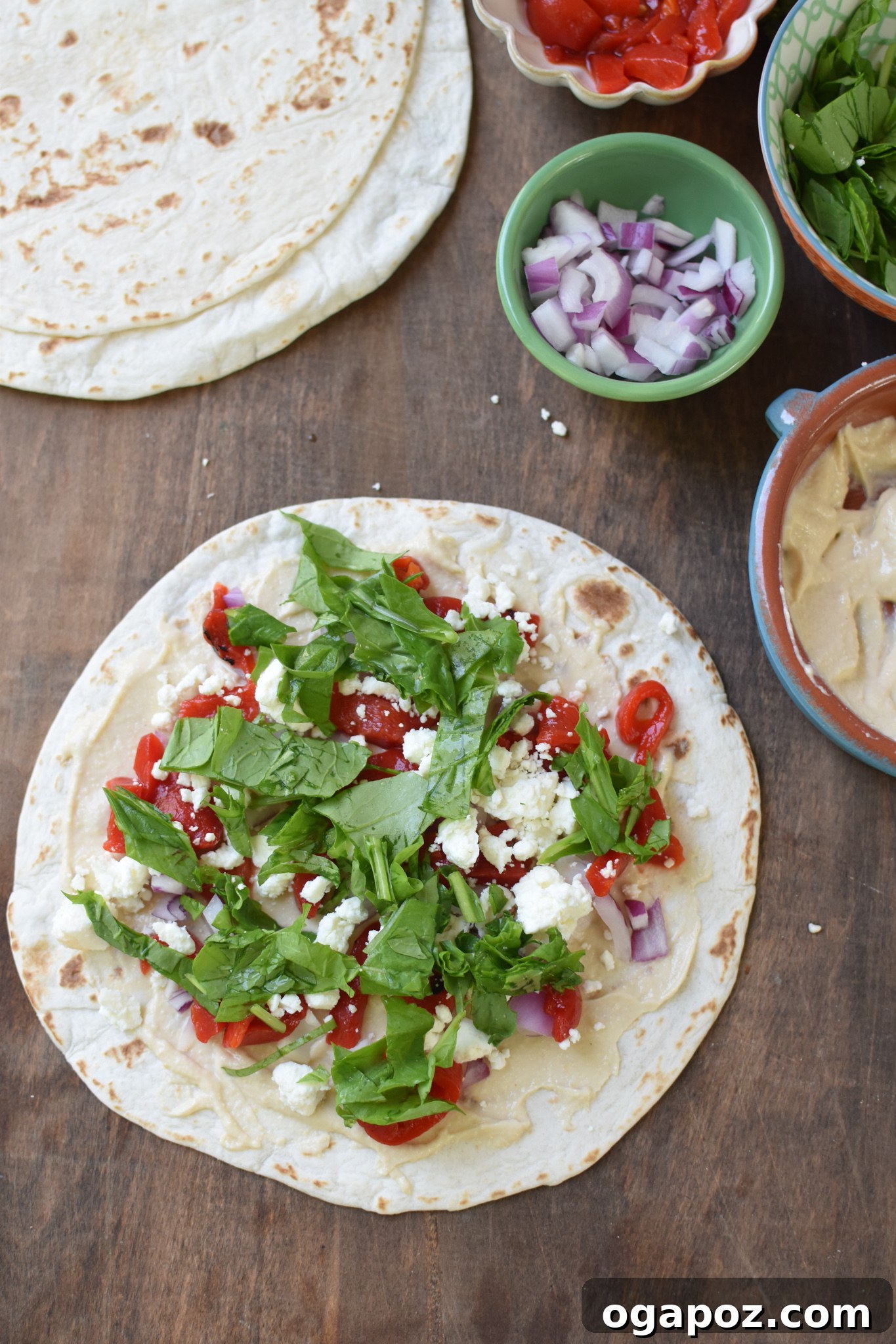 A tortilla layered with hummus and an assortment of Mediterranean ingredients including roasted red peppers, feta, arugula, red onion, and parsley.