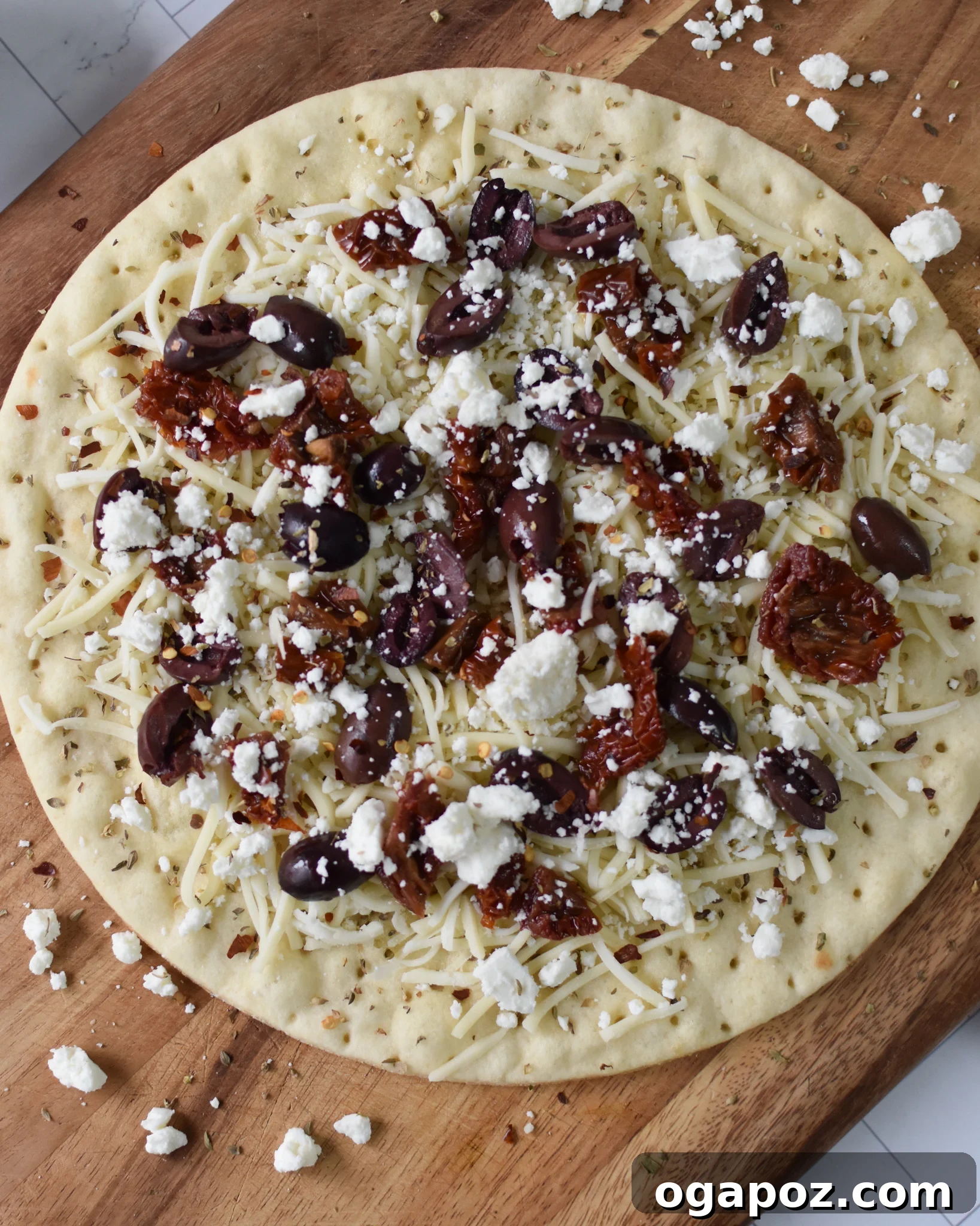 Aegean Delight Pizza 5 Unbaked Greek Pizza with cheese, sun-dried tomatoes, and olives visible on the dough.