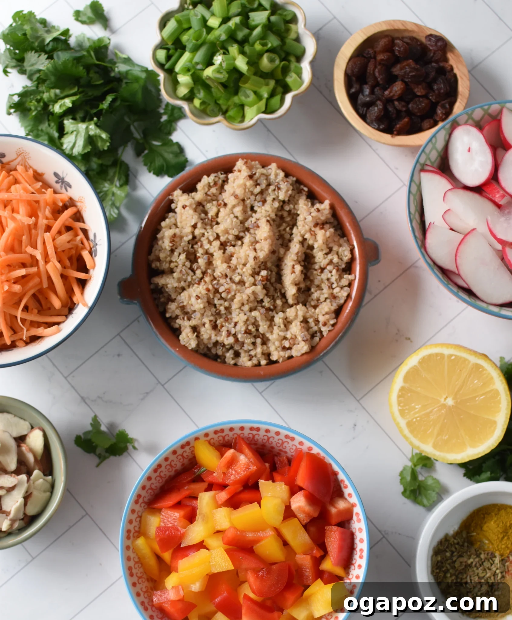 A flat lay photograph displaying all the fresh ingredients for Curried Quinoa Salad laid out on a wooden surface: cooked quinoa, sliced carrots, bell peppers, radishes, raisins, almonds, cilantro, green onions, and various dressing components.