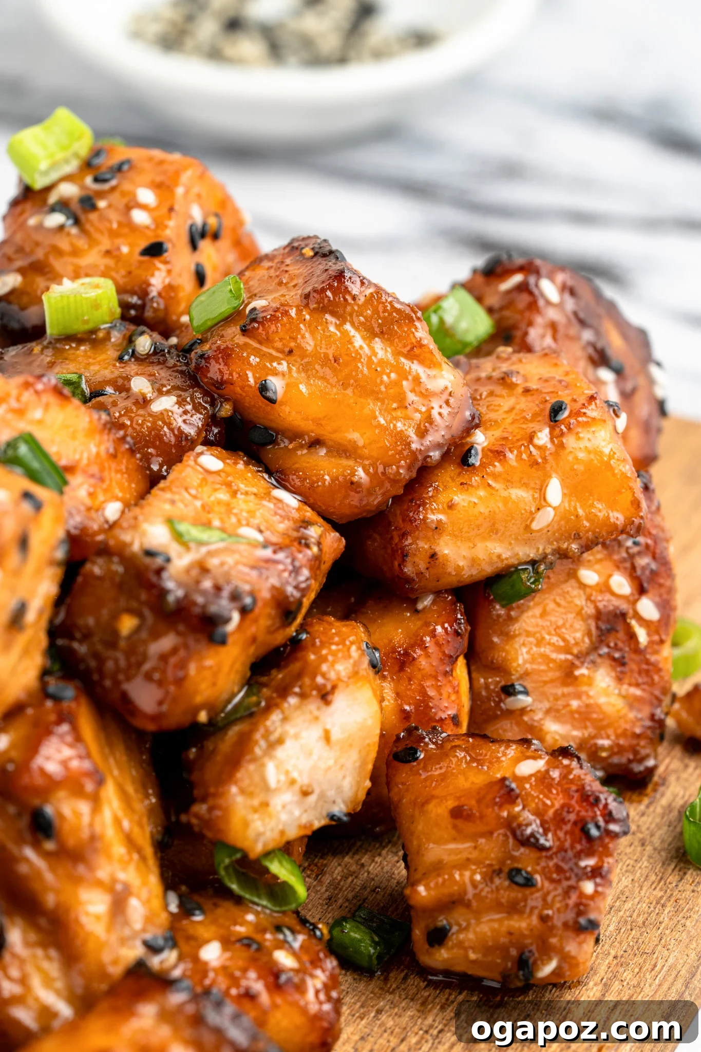 Crispy Air-Fried Teriyaki Salmon Poppers 2 Close-up of glazed salmon cubes garnished with sesame seeds and green onions on a wooden board. A small bowl of sesame seeds is blurred in the background.
