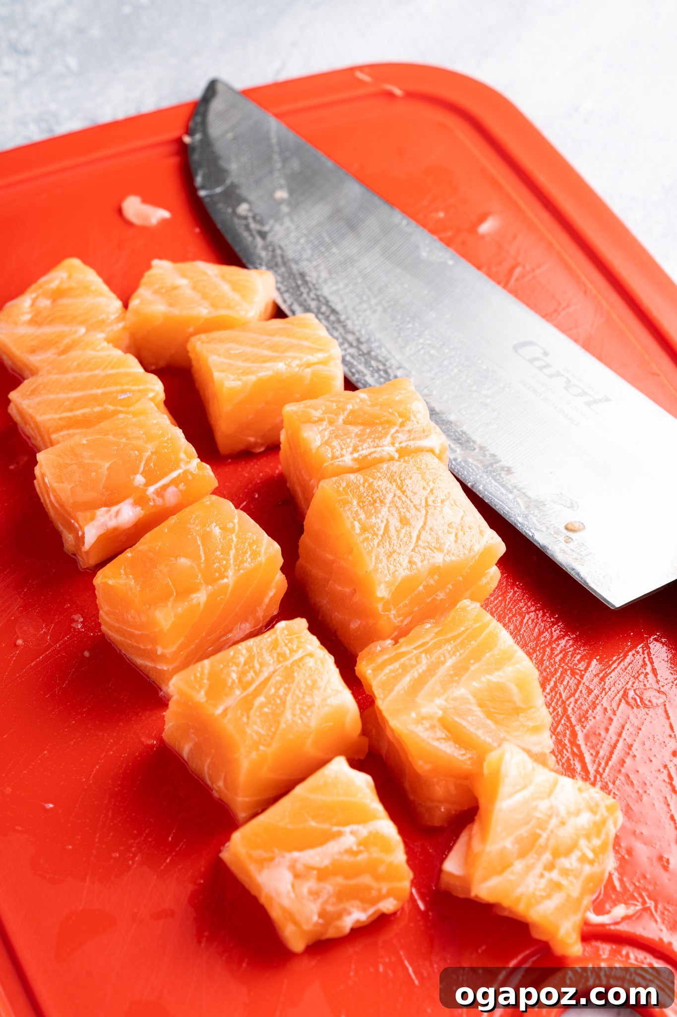 Crispy Air-Fried Teriyaki Salmon Poppers 6 Cubes of fresh salmon are neatly arranged on a bright red cutting board. A large kitchen knife with a shiny blade is positioned next to the salmon pieces. The background has a textured, cool-toned surface.