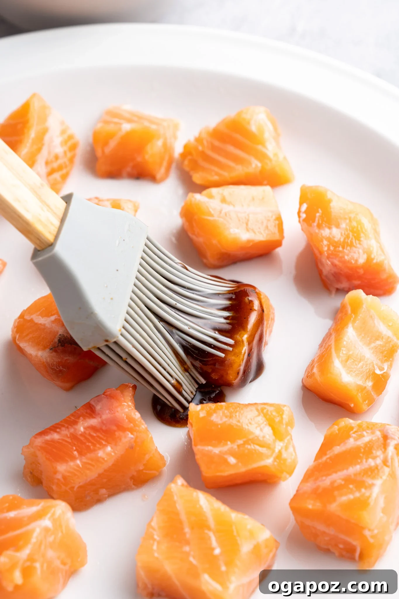 Crispy Air-Fried Teriyaki Salmon Poppers 7 A silicone brush applies a dark marinade to raw salmon cubes on a white plate. The salmon pieces are evenly spaced and glistening, suggesting preparation for cooking. The background is softly blurred.