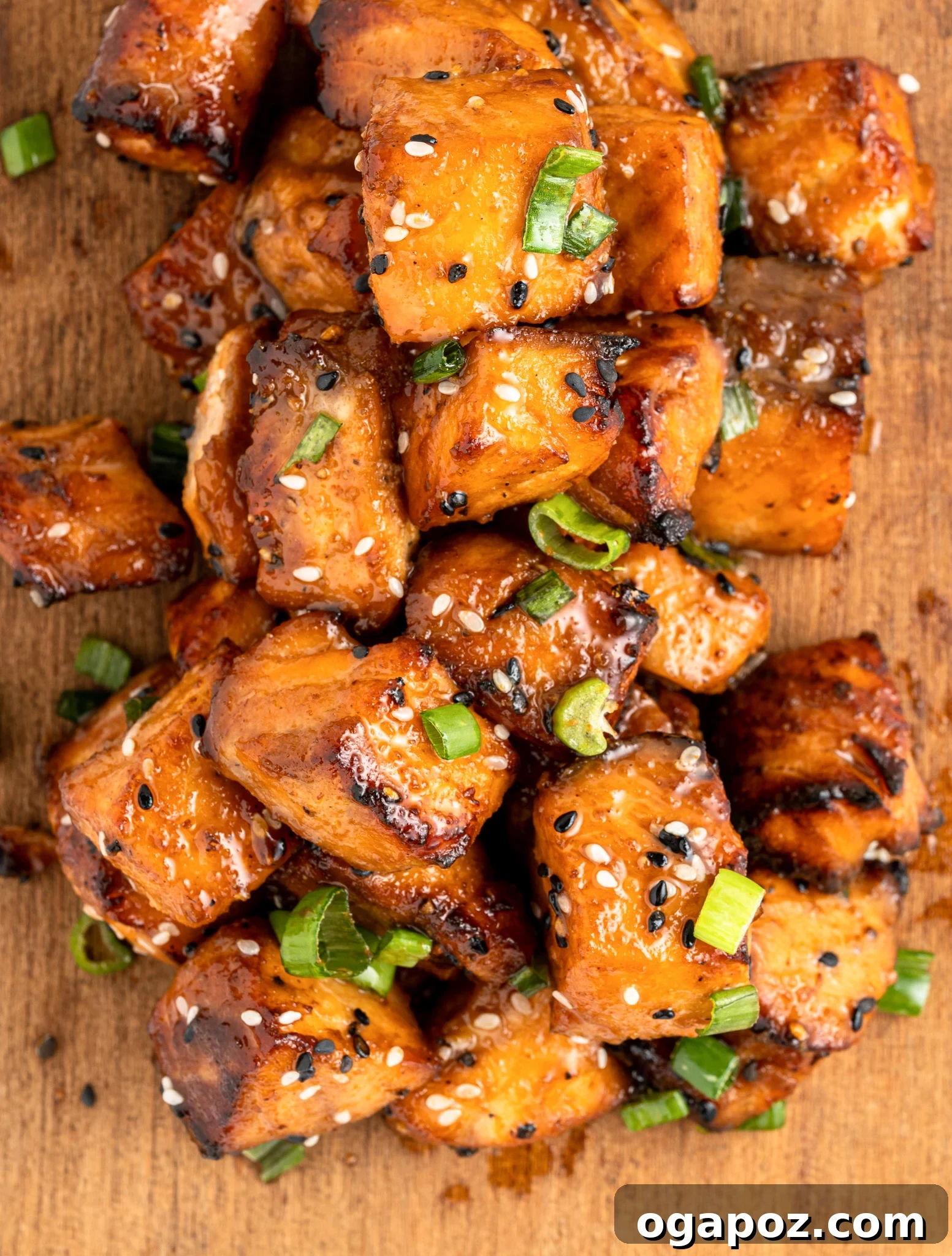 Crispy Air-Fried Teriyaki Salmon Poppers 9 A pile of glazed, air fryer salmon bites topped with sesame seeds and sliced green onions on a wooden surface. The salmon has a caramelized appearance, indicating its well-cooked and flavorful.