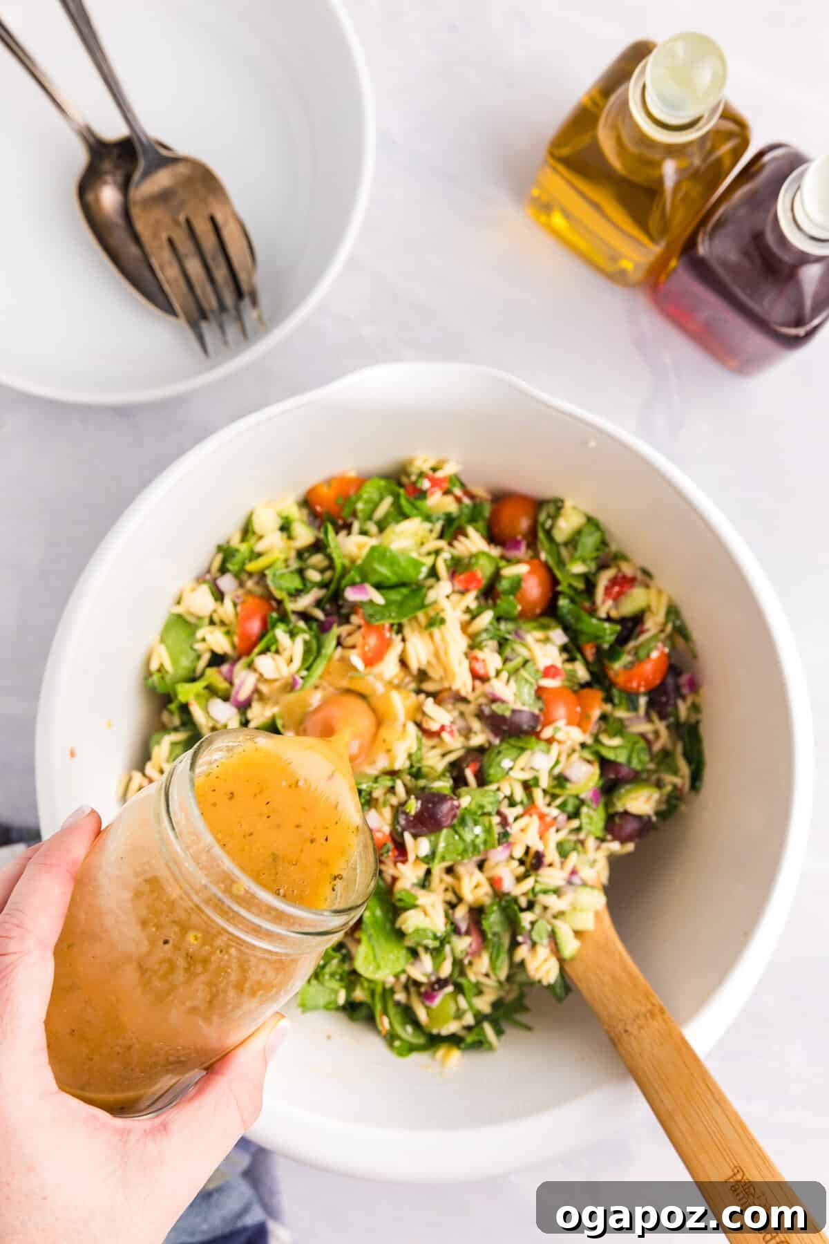 Mediterranean Garden Pasta 10 A generous stream of vibrant homemade Greek dressing being poured over the Mediterranean Pasta Salad in a large mixing bowl, enhancing its flavors.