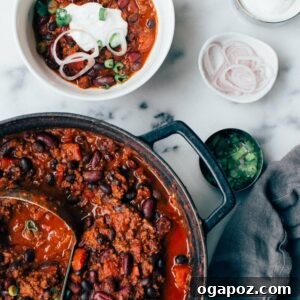 Ground beef chili in a pot with a bowl of chili next to it, topped with cheese and sour cream.