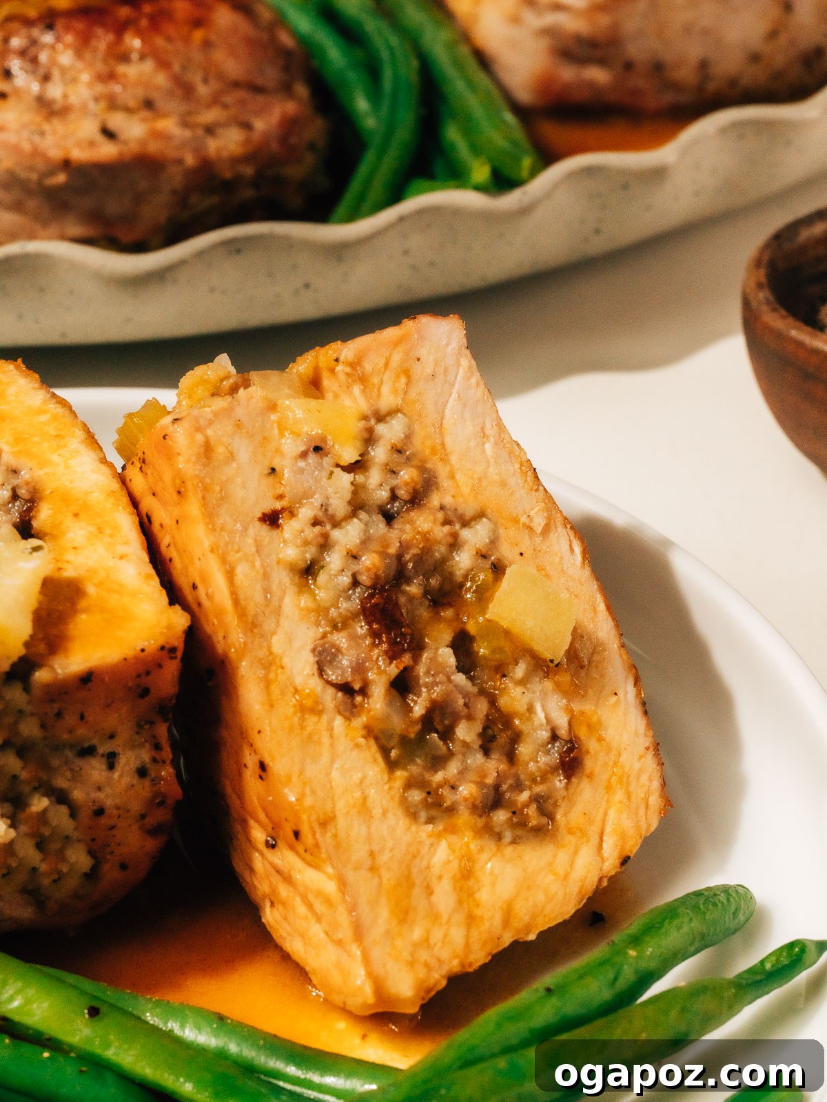 Signature Stuffed Pork Chops with an Unforgettable Pan Sauce 2 A perfectly seared and stuffed pork chop on a plate, cut open to reveal the delicious sausage-apple filling. More stuffed pork chops are blurred in the background, suggesting a generous meal.