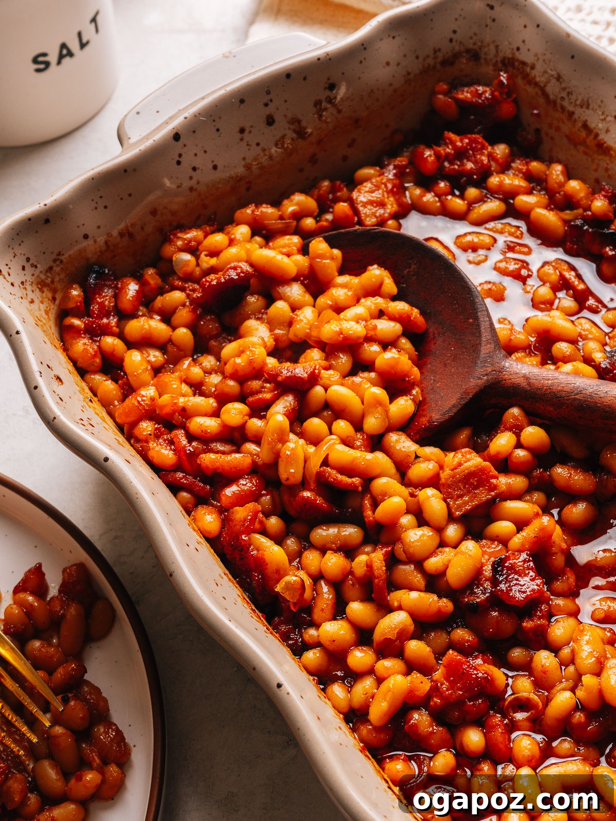 Baked beans in a baking dish with a wooden spoon and a plated serving alongside.