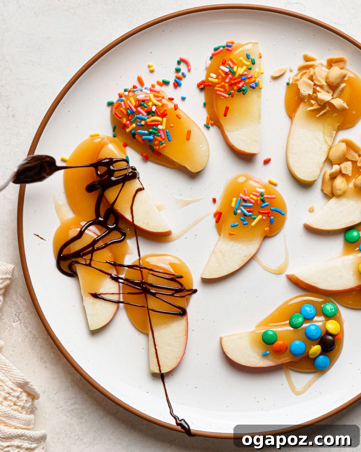 Caramel dipped apple slices with sprinkles, peanuts and mini M&M’s on a white plate.